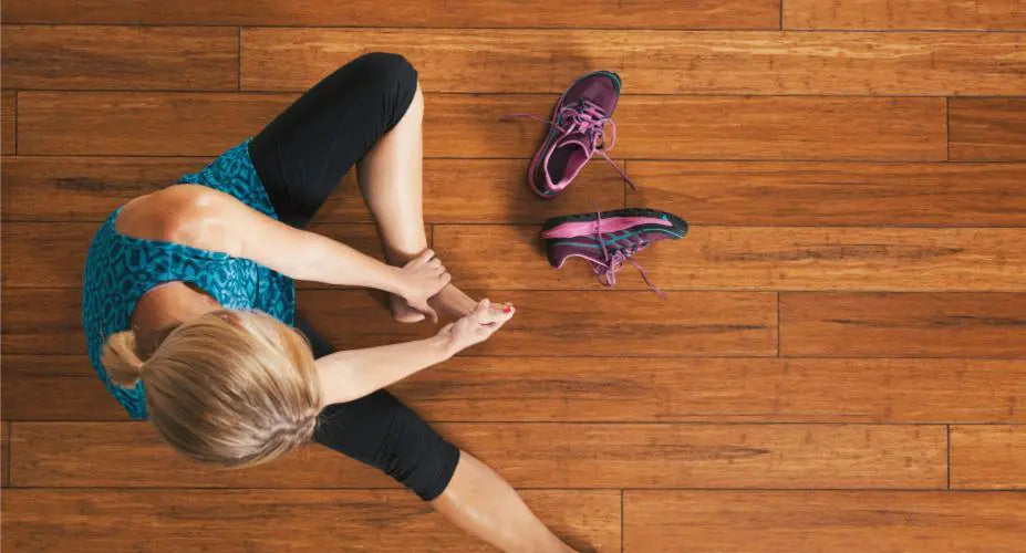 A woman in athletic clothing sits on a wooden floor stretching her legs, with a pair of purple running shoes placed nearby.